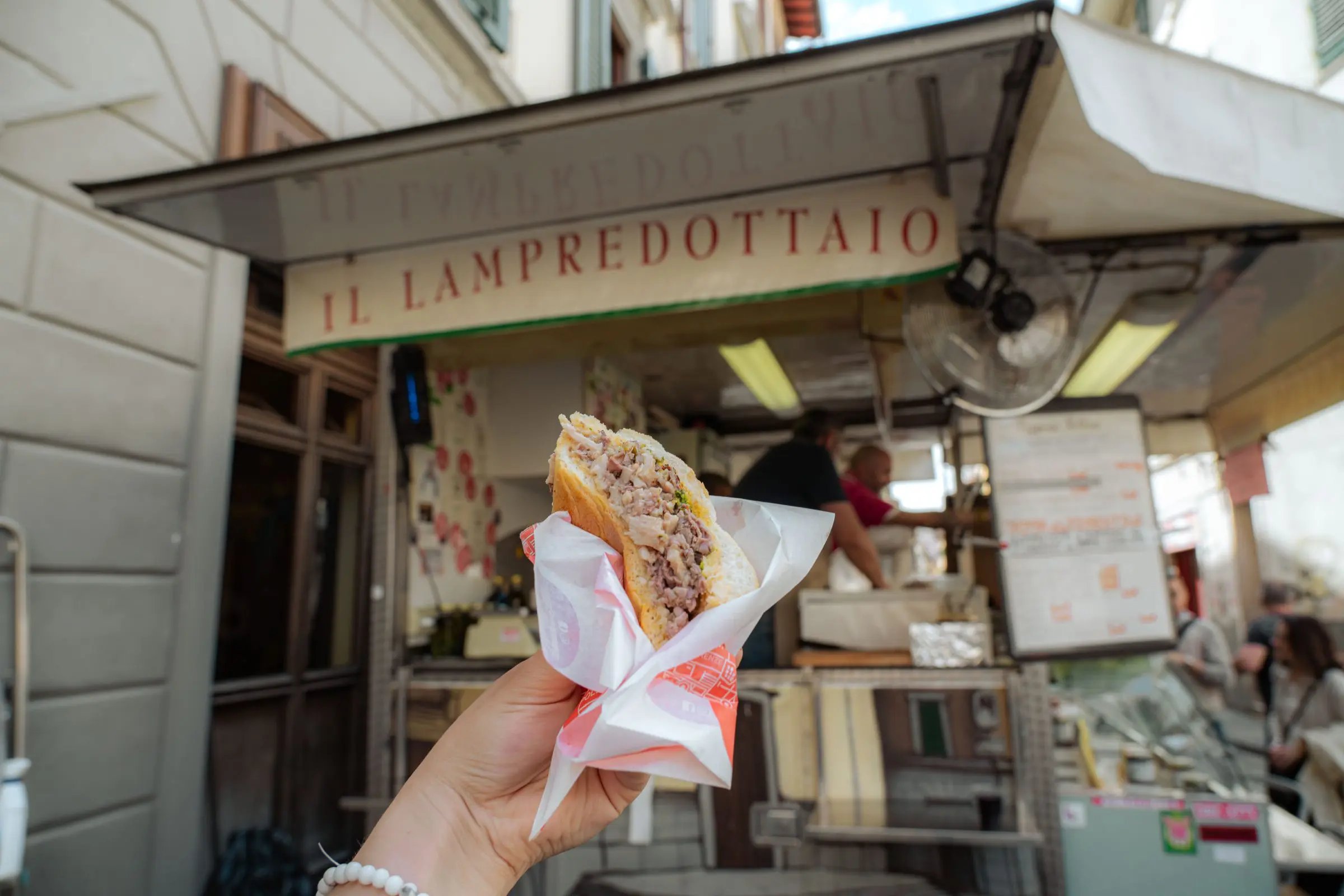 A hand is holding a lampredotto sandwich in front of a lampredotto food truck.