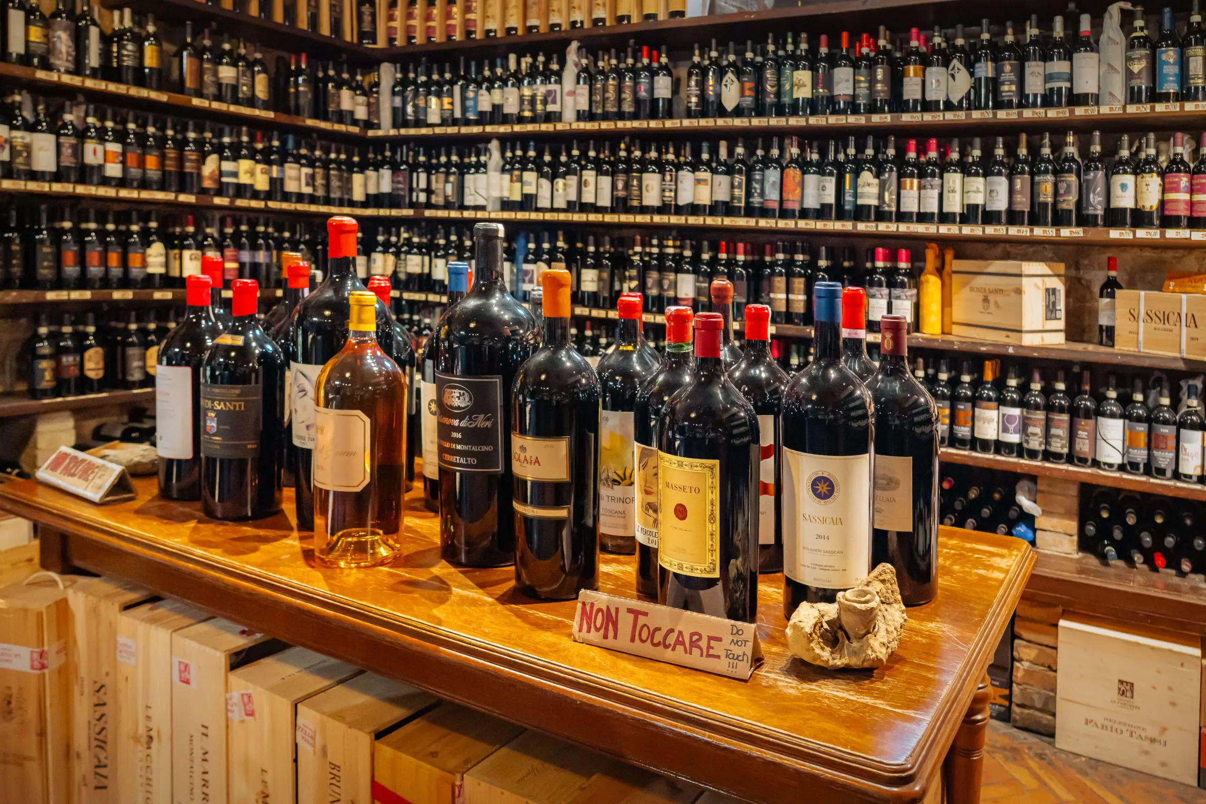 A wooden table with many bottles of wine on it and shelves filled with wine bottles in the background.