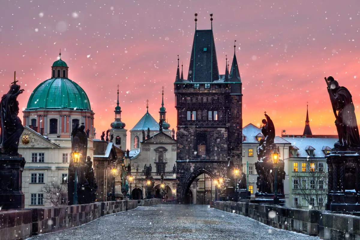 Sunrise over the Charles Bridge in Prague. The sky is pink and it's snowing.
