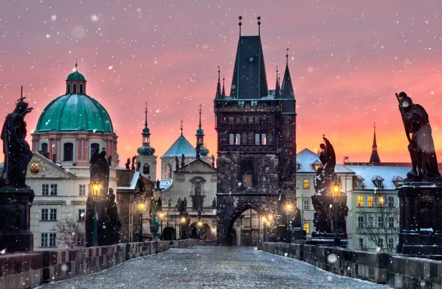 Sunrise over the Charles Bridge in Prague. The sky is pink and it's snowing.