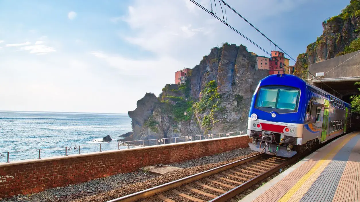 A train traveling along the coast with a colorful village atop a hill in the background. 