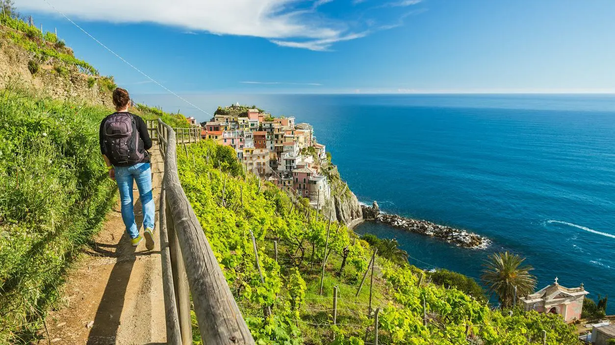 A hiker walking along a narrow path lined with vineyards, enjoying the view of Manarola and the sea. 