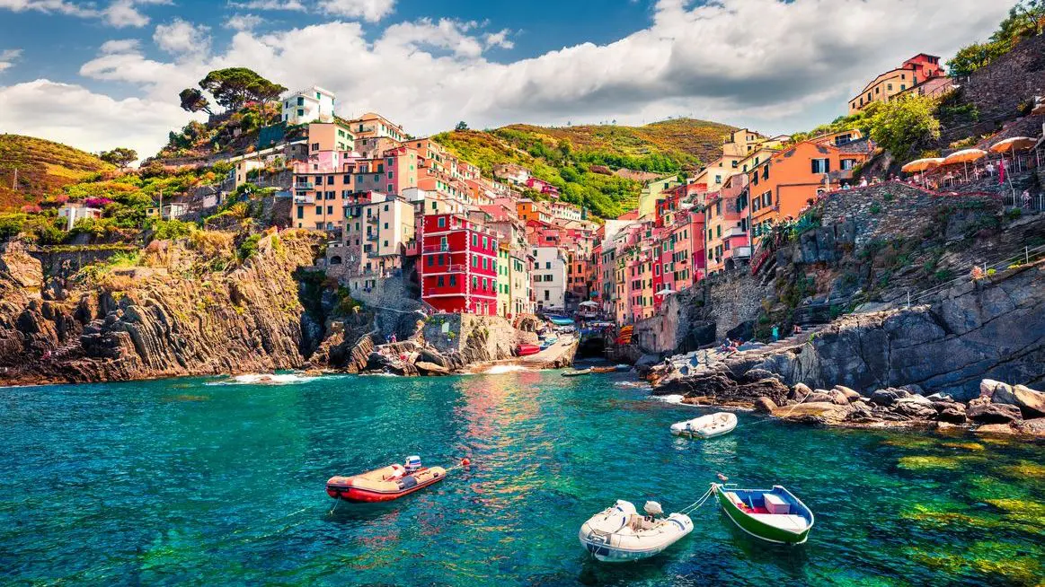 A panoramic view of Riomaggiore and its colorful buildings perched atop steep hillsides. The emerald sea is in the foreground. 
