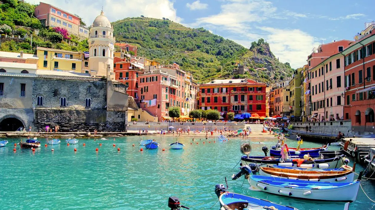 A panoramic view of the colorful buildings of Vernazza, with hilltop terraced vineyards in the background and small wooden boats on turquoise waters in the foreground. 