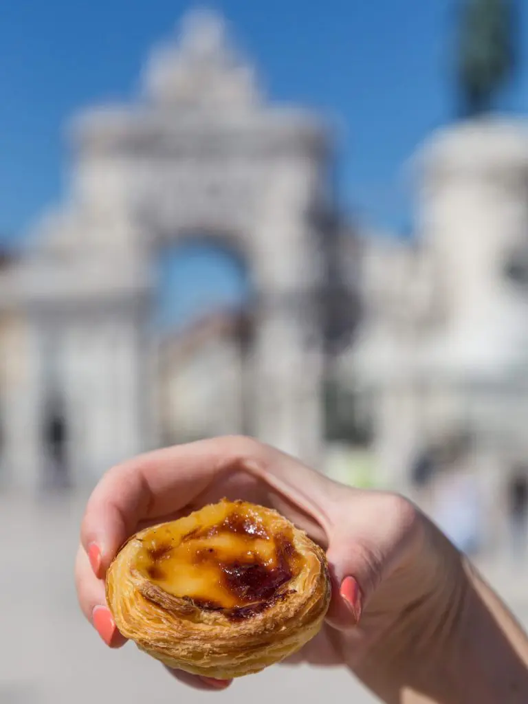 Traditional Portuguese egg tart pasty cake dessert Pasteis de nata in women hand. On background attractions in Lisbon, Portugal.