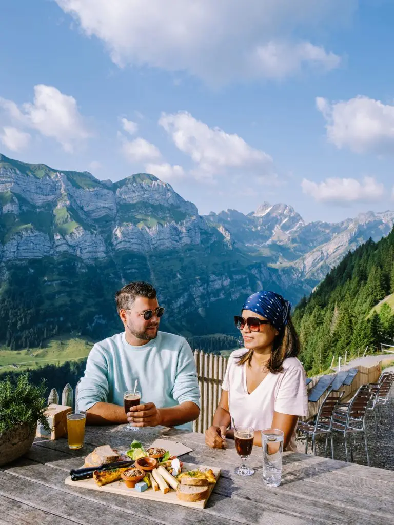 A couple enjoying drinks and traditional Swiss food at an outdoor mountain restaurant in the Swiss Alps, with panoramic alpine peaks and green valleys in the background.
