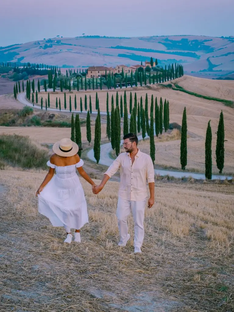 A couple holding hands while walking through rolling hills in Tuscany, Italy, with a winding cypress-lined road and a countryside villa in the background at sunset.