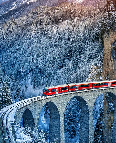 A bright red train crossing a curved stone viaduct in the snowy Swiss Alps, surrounded by dense pine forests and steep mountain cliffs dusted with winter snow.