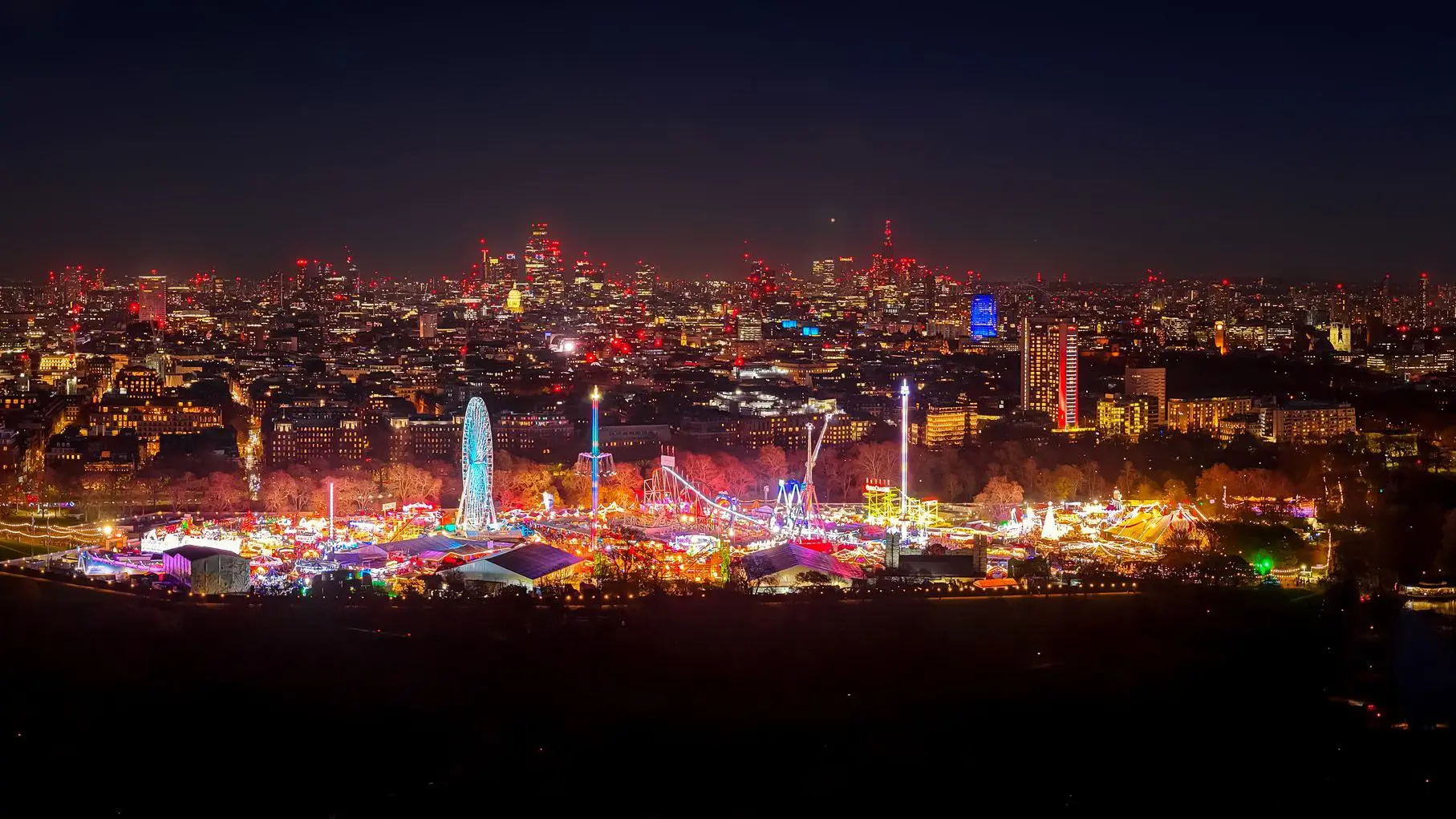 Vibrant Christmas fair lights glow in Hyde Park beneath the London skyline at dusk, featuring rides, a Ferris wheel, and festive cheer.
