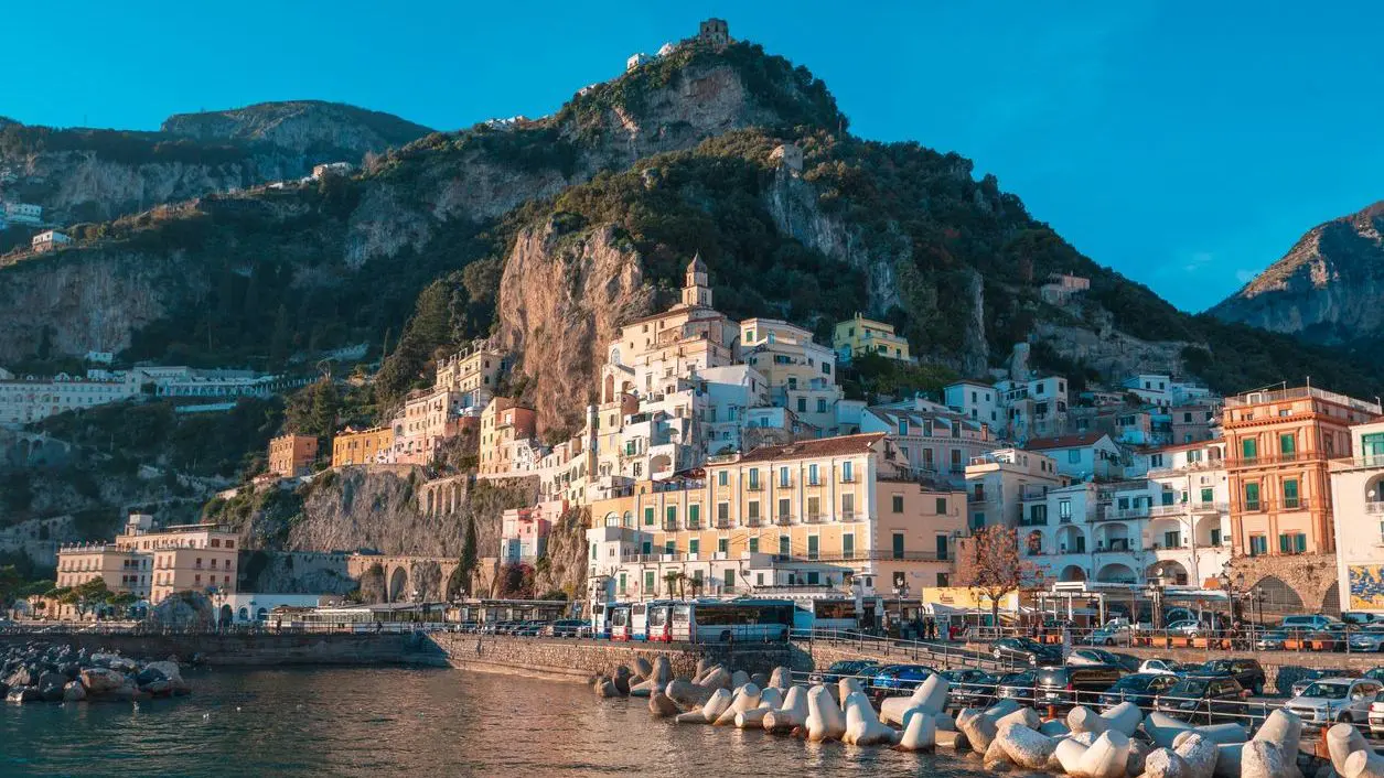 The picturesque seaside town of Amalfi in Italy nestled against rocky cliffs with lush greenery, featuring colorful buildings, a moored boat, and a clear blue sky.