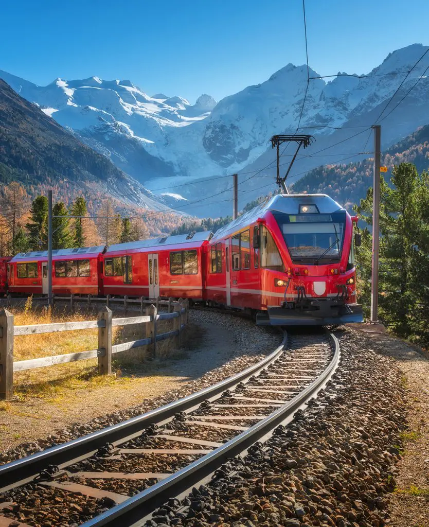 The red Bernina Express train rounds a curve through an autumn Alpine landscape, with golden larch trees, evergreen pines, and dramatic snow-capped peaks and a glacier visible under a clear blue sky.