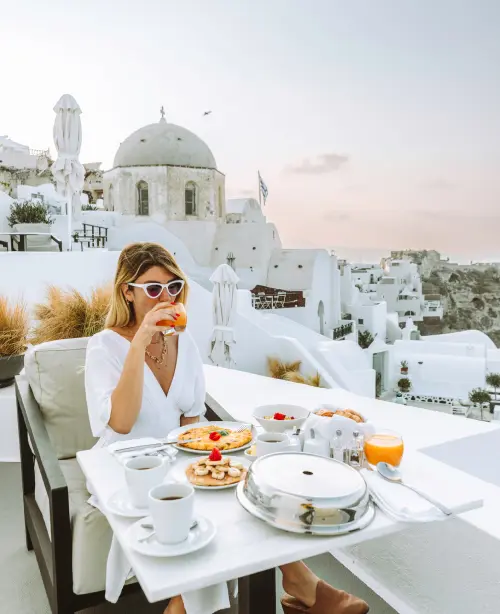 Woman enjoying a drink in Oia with whitewashed buildings and Aegean Sea views in the background.