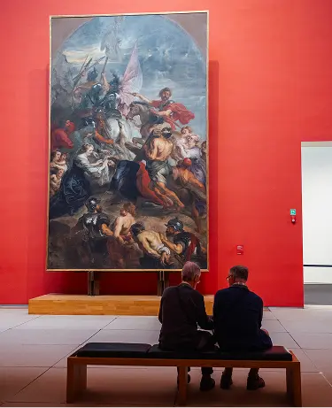 Two visitors seated on a bench contemplating a large Baroque painting in a red-walled museum gallery
