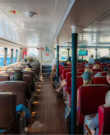 Passengers seated in red chairs inside the cabin of a ferry, bags stowed beneath seats, with a staircase leading to the upper deck visible ahead.