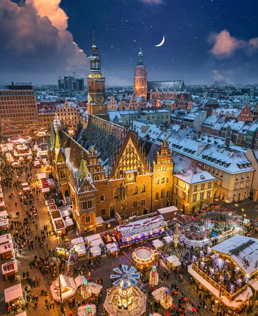 Festive European Christmas market with illuminated wooden stalls, holiday decorations, and crowds browsing seasonal gifts at night.