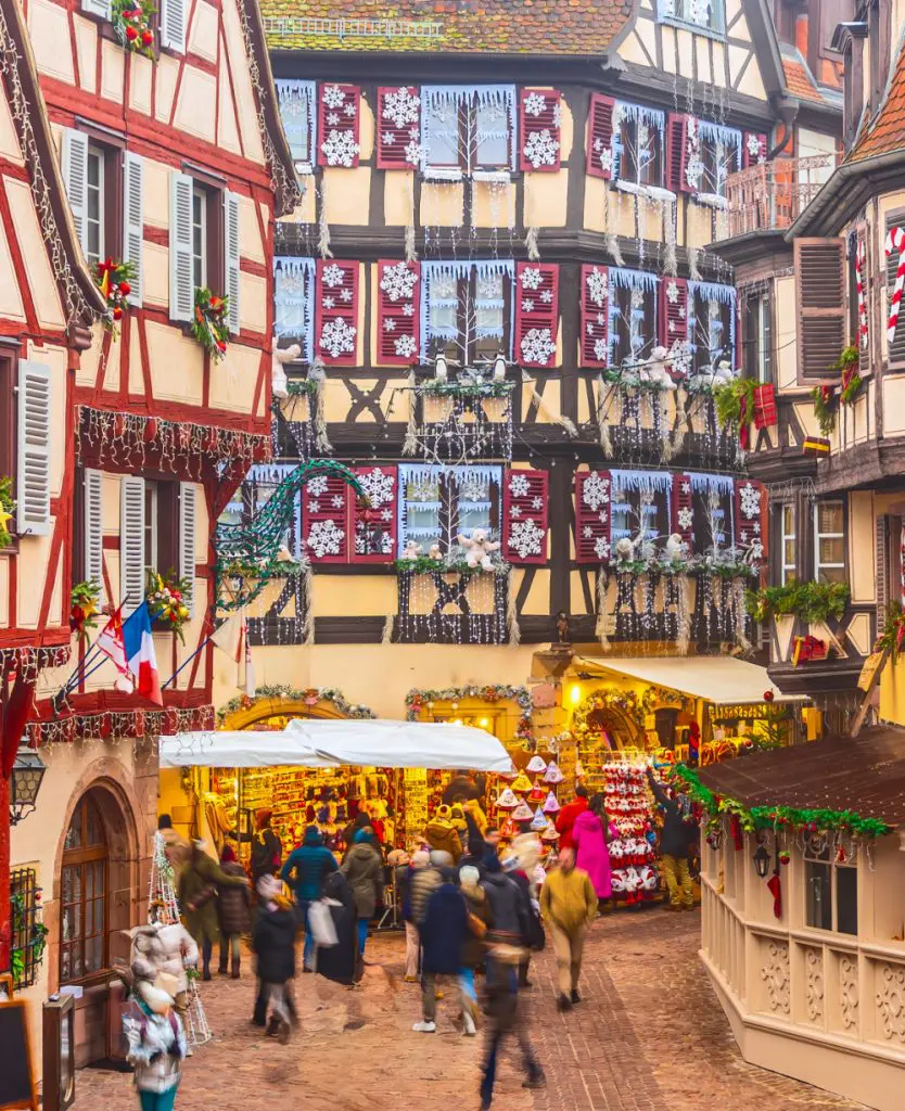Festive European Christmas market with illuminated wooden stalls, holiday decorations, and crowds browsing seasonal gifts at night.