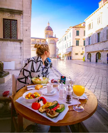 A woman dining al fresco at a cafe on Dubrovnik's Stradun, with the cathedral dome and limestone old town in the background