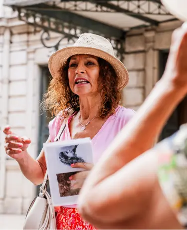 A female tour guide in a straw hat enthusiastically explaining a point of interest to guests during a city walking tour