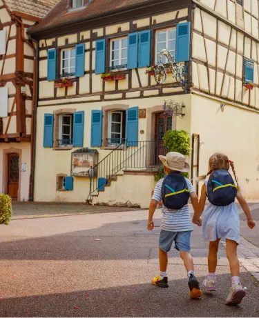 Two young children wearing small backpacks walk hand in hand toward a traditional Alsatian half-timbered house with blue shutters and flower-filled windowboxes.