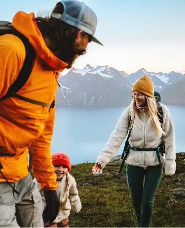 A family of three hiking in cool-weather gear pause on a mountain trail, the parents smiling at their toddler, with a dramatic fjord and snow-dusted peaks stretching into the distance behind them.