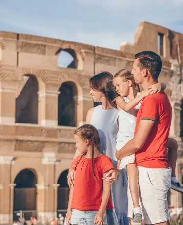 A family of four — two parents and two young girls in matching red tops — stand together gazing up at the ancient arches of the Colosseum in Rome on a sunny day.