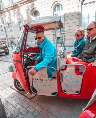 A guide driving two tourists through the streets of Budapest in a red tuk-tuk displaying the Budapest Card