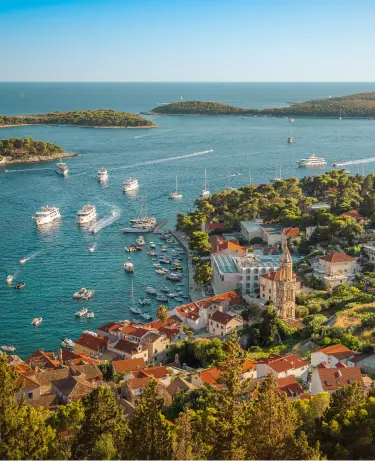 Aerial view of Hvar town on Croatia's Dalmatian coast, with terracotta-roofed stone buildings and a church tower in the foreground, yachts and cruise boats dotting the sparkling turquoise bay, and forested islands stretching into the distance.