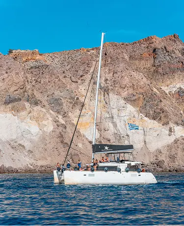 A white catamaran with passengers on deck anchored in deep blue water off the dramatic volcanic cliffs of Santorini, with a Greek flag painted on the rocky face behind the boat.