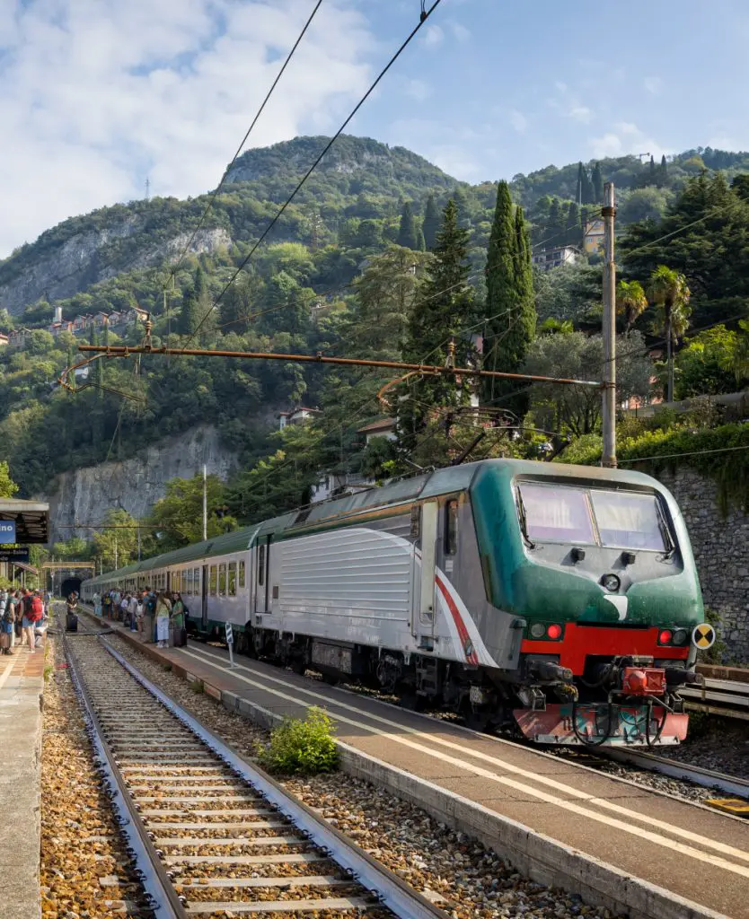 A green and silver Trenitalia locomotive pulls passenger carriages into a small station on the Lake Como line, with travellers waiting on the platform and lush tree-covered hillsides and terracotta-roofed villas rising steeply behind.