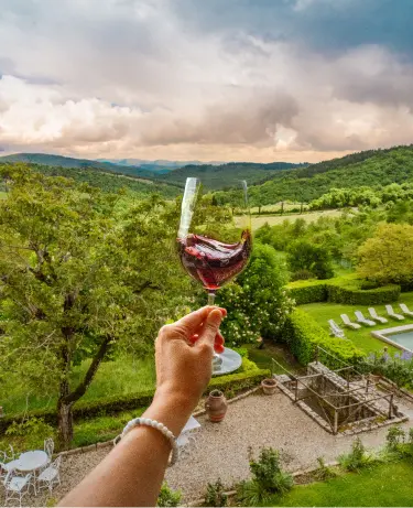 A hand holding a glass of red wine over a lush Tuscan valley with rolling hills and a villa garden below