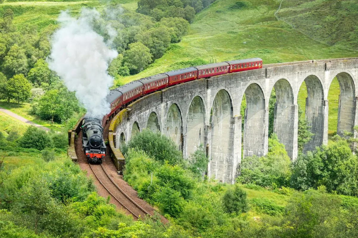 The iconic red Jacobite steam train traveling along the Glenfinnan Viaduct amidst lush greenery.