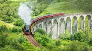 The iconic red Jacobite steam train traveling along the Glenfinnan Viaduct amidst lush greenery.