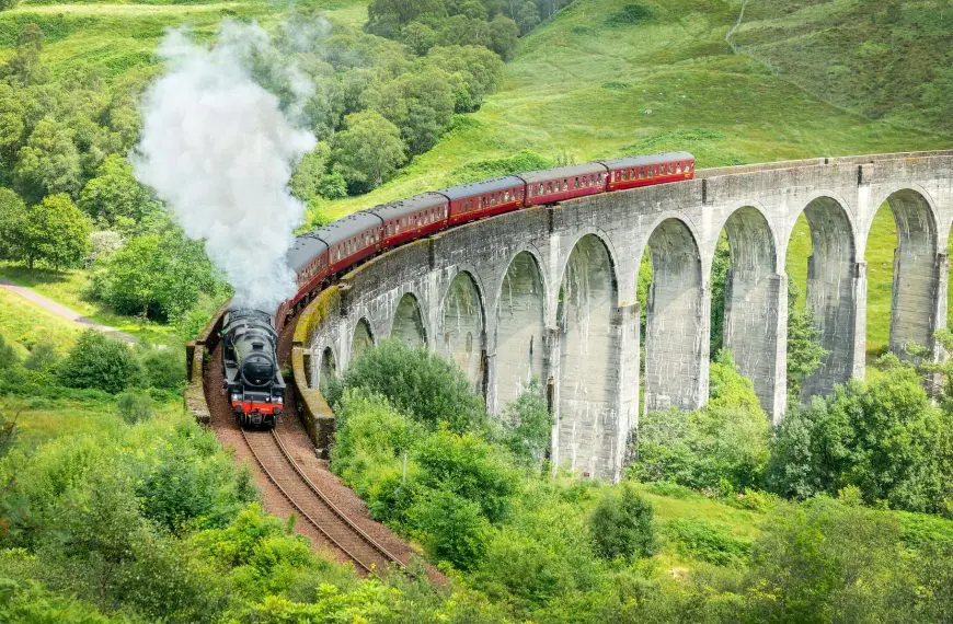 The iconic red Jacobite steam train traveling along the Glenfinnan Viaduct amidst lush greenery.