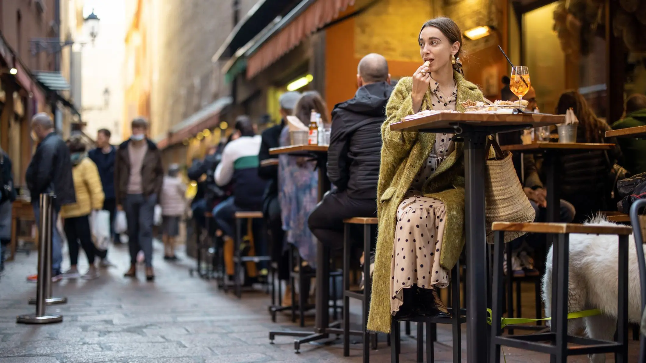 Una mujer sentada en un bar al aire libre en una calle concurrida, comiendo bocadillos y bebiendo un Aperol Spritz. 