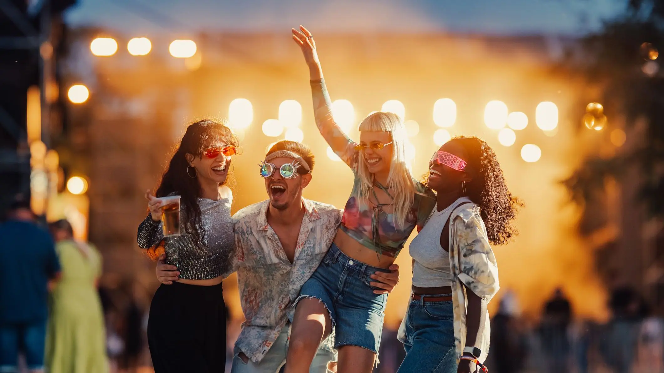 Four friends are laughing and having fun at an outdoor music festival, enjoying a live concert.