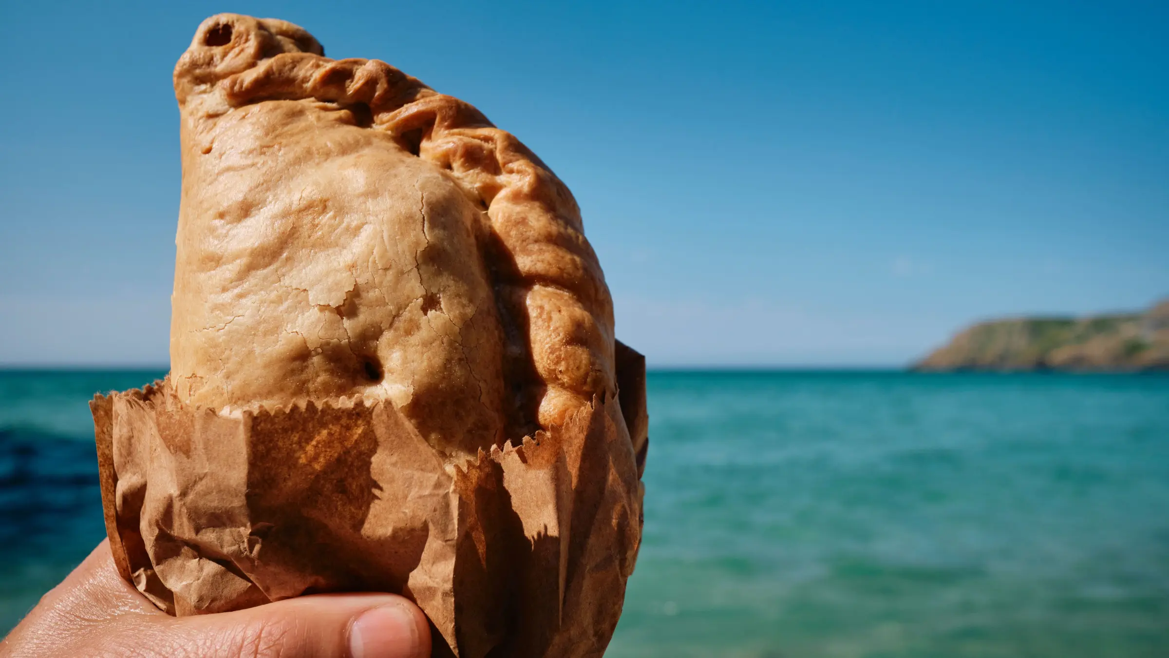 A hand holding a Cornish pasty with the sea in the background.
