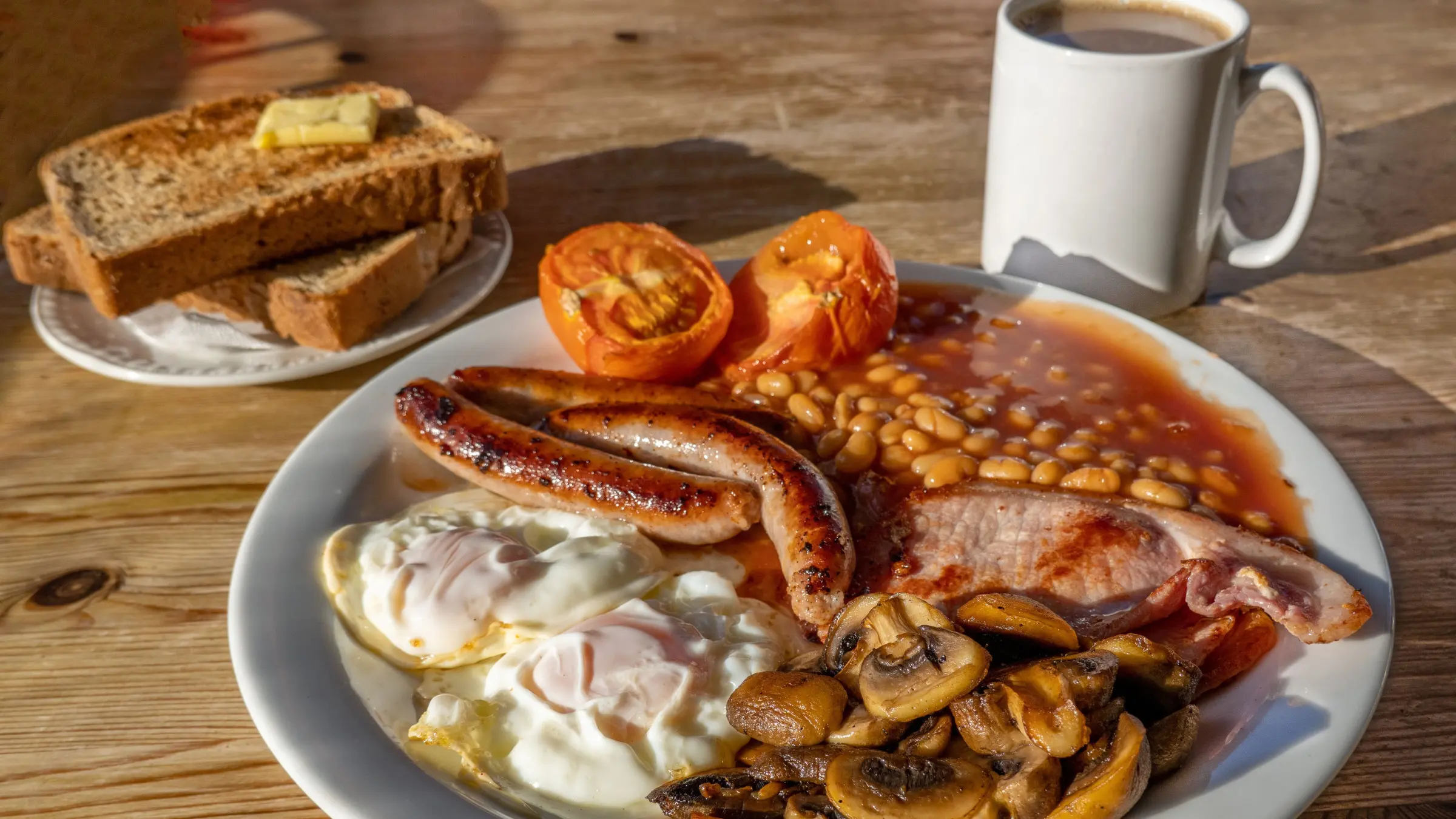 A close-up of a full English breakfast with fried eggs, sausages, bacon, tomatoes, beans, and mushrooms. There's also a plate with toasted bread and butter and a cup of coffee.