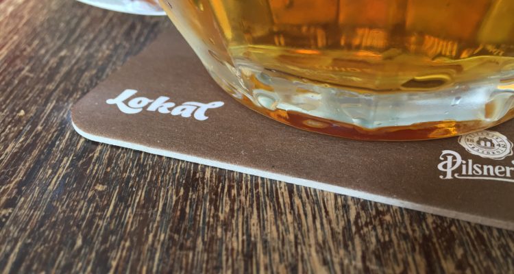 A close-up photo of a glass of beer on a wooden table, resting on a branded brown beer coaster