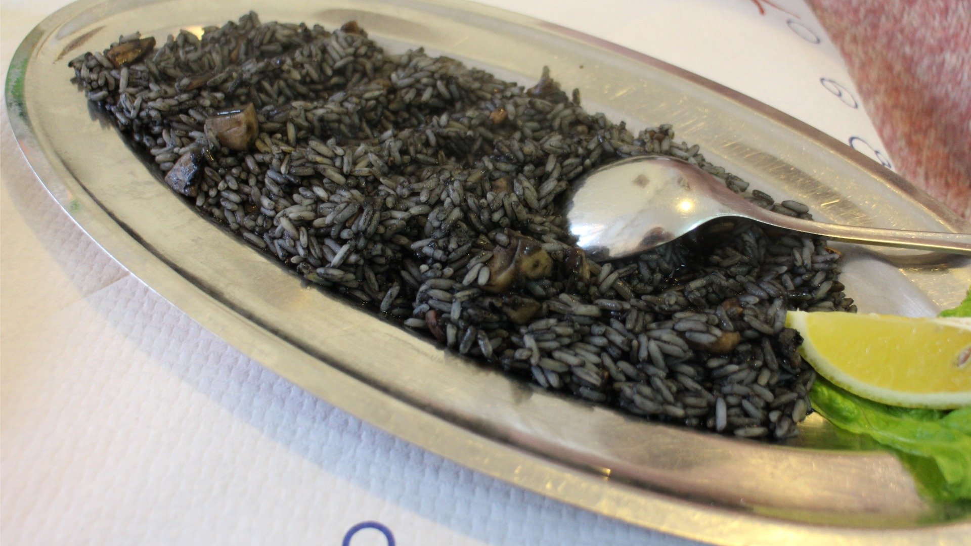 A stainless steel serving platter filled with black-colored rice and a lemon wedge on the side. 