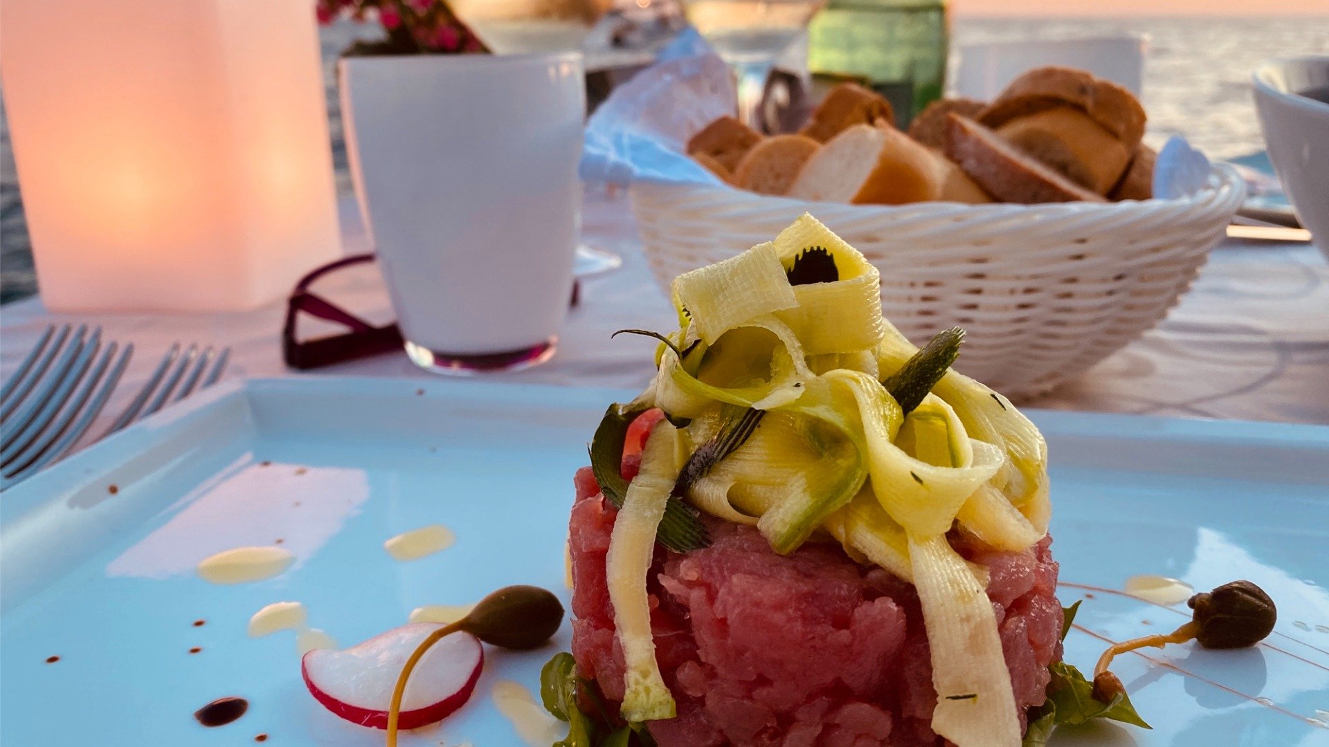 A tuna tartare topped with zucchini strips and a bread basket in the background. 