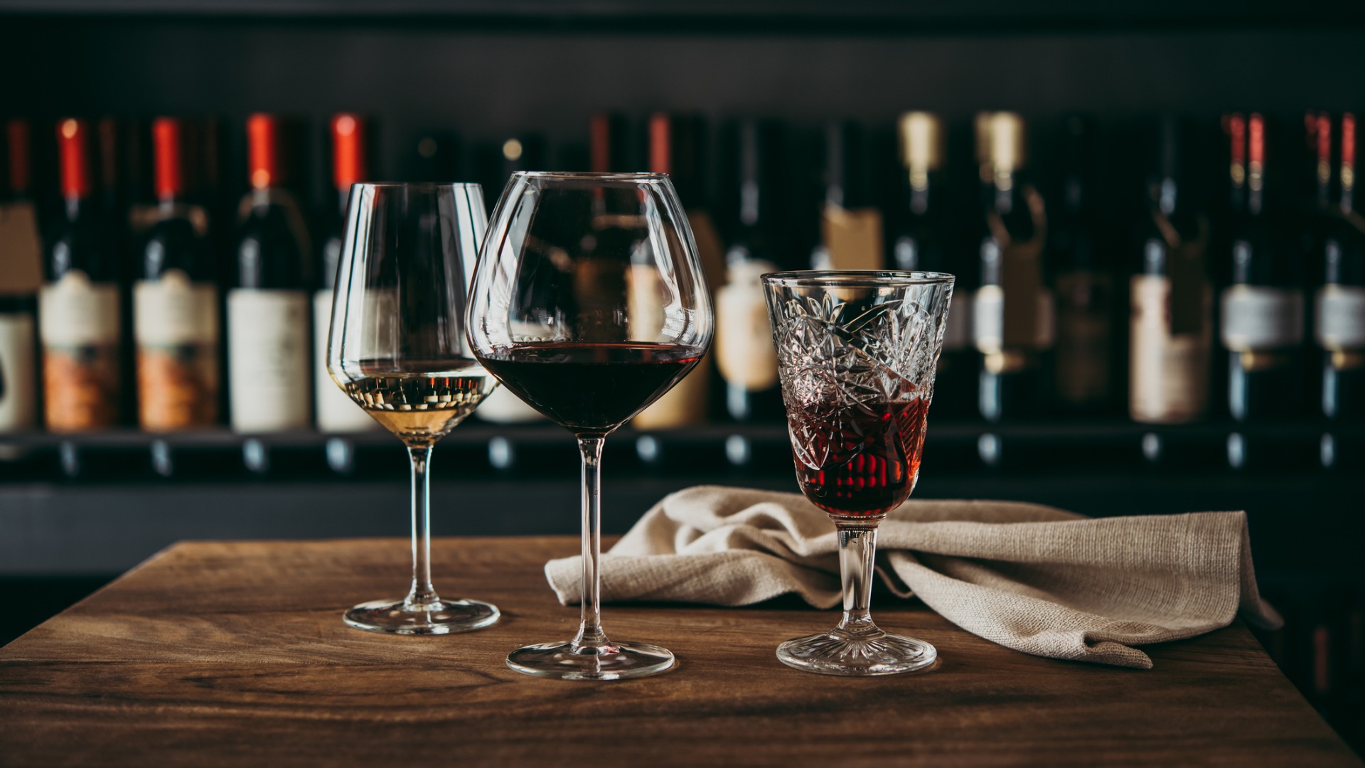 Această imagine prezintă trei pahare diferite de vin umplute cu vin alb, roșu și, respectiv, rose.  Pe fundal un perete plin cu sticle de vin. 