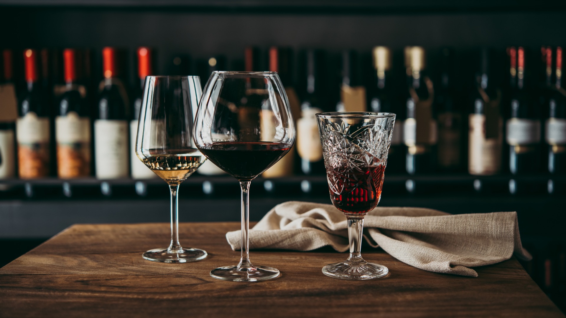 This image shows three different glasses of wine filled with white, red, and rose wine respectively. In the background a wall filled with bottles of wine. 