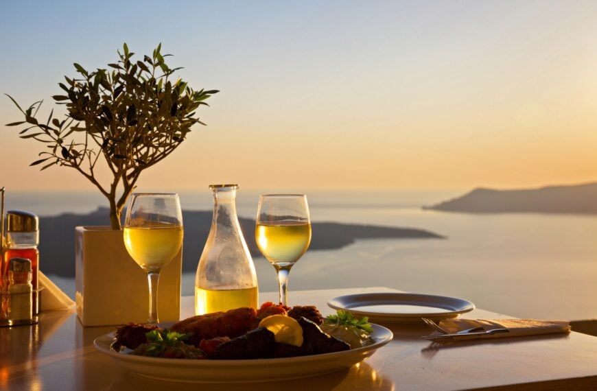 This image shows a table set for two at sunset in Santorini. There's a carafe and two glasses of white wine, and a platter with Greek food.