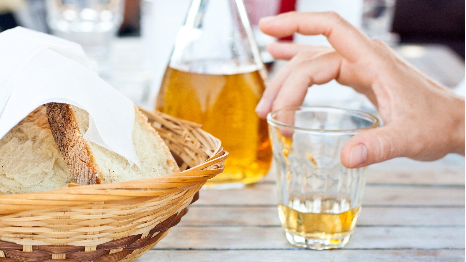 This is a close up of a basket filled with bread and someone's hand holding a glass of Greek retsina wine. In the background, a carafe of retsina. 