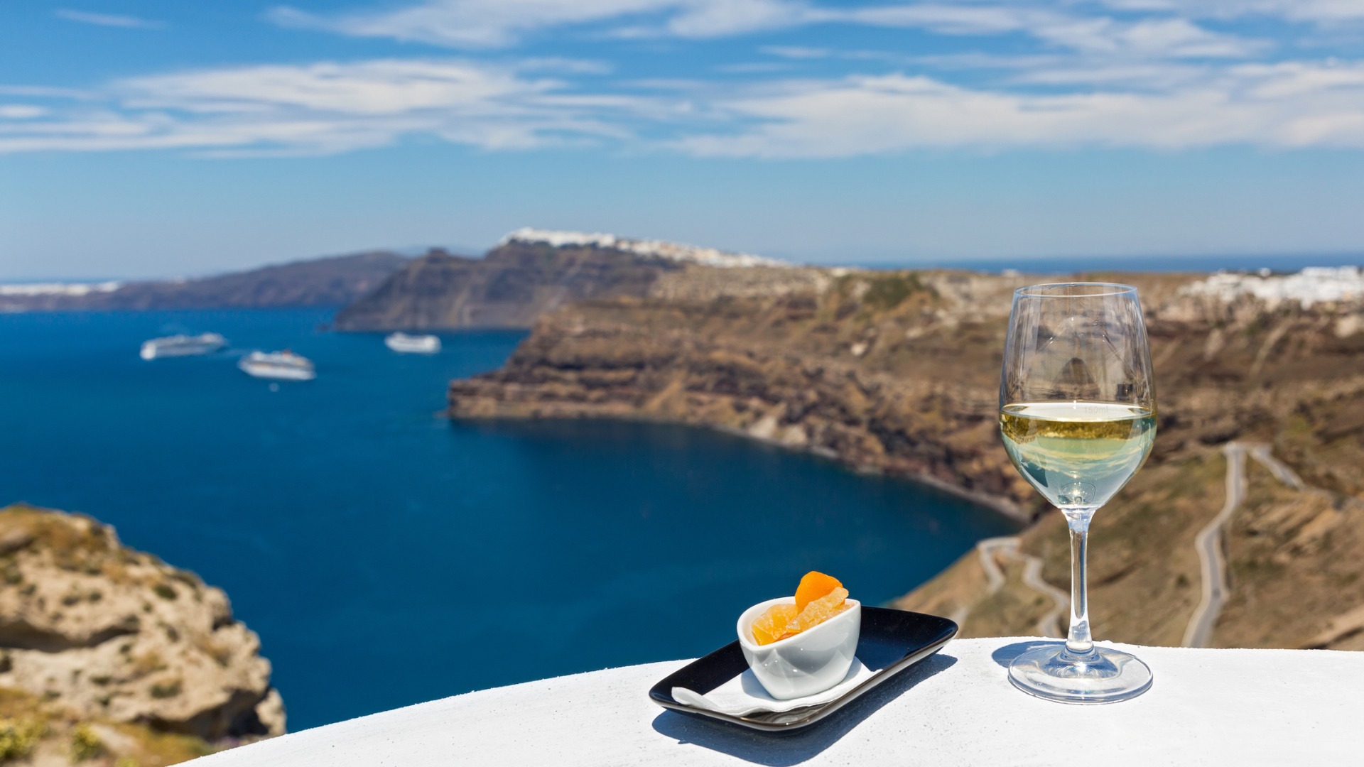 Această imagine arată un pahar de vin alb în prim plan, cu caldera din Santorini în fundal.  Santorini produce unele dintre cele mai bune vinuri grecești. 