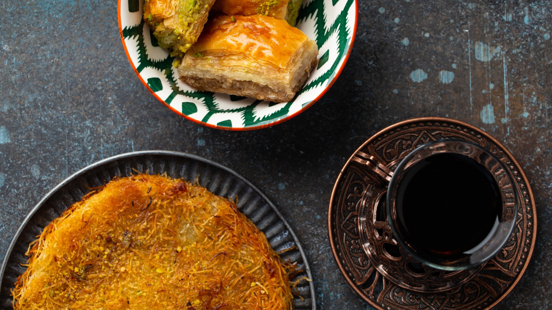This is a top-down photo that shows a kunefe dessert, some pieces of baklava and a glass of traditional Turkish tea. 