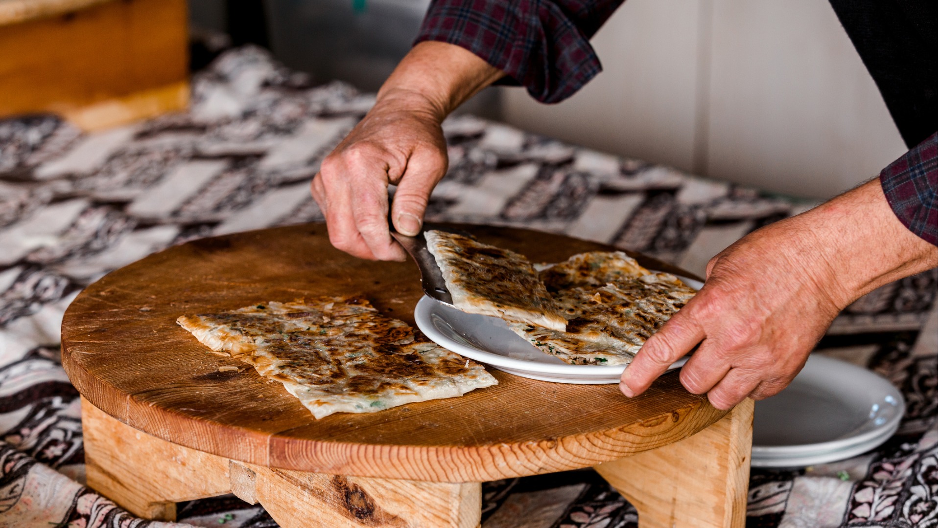 This is a close-up of two hands cutting gozleme on a wooden board and serving the pieces on a plate. 