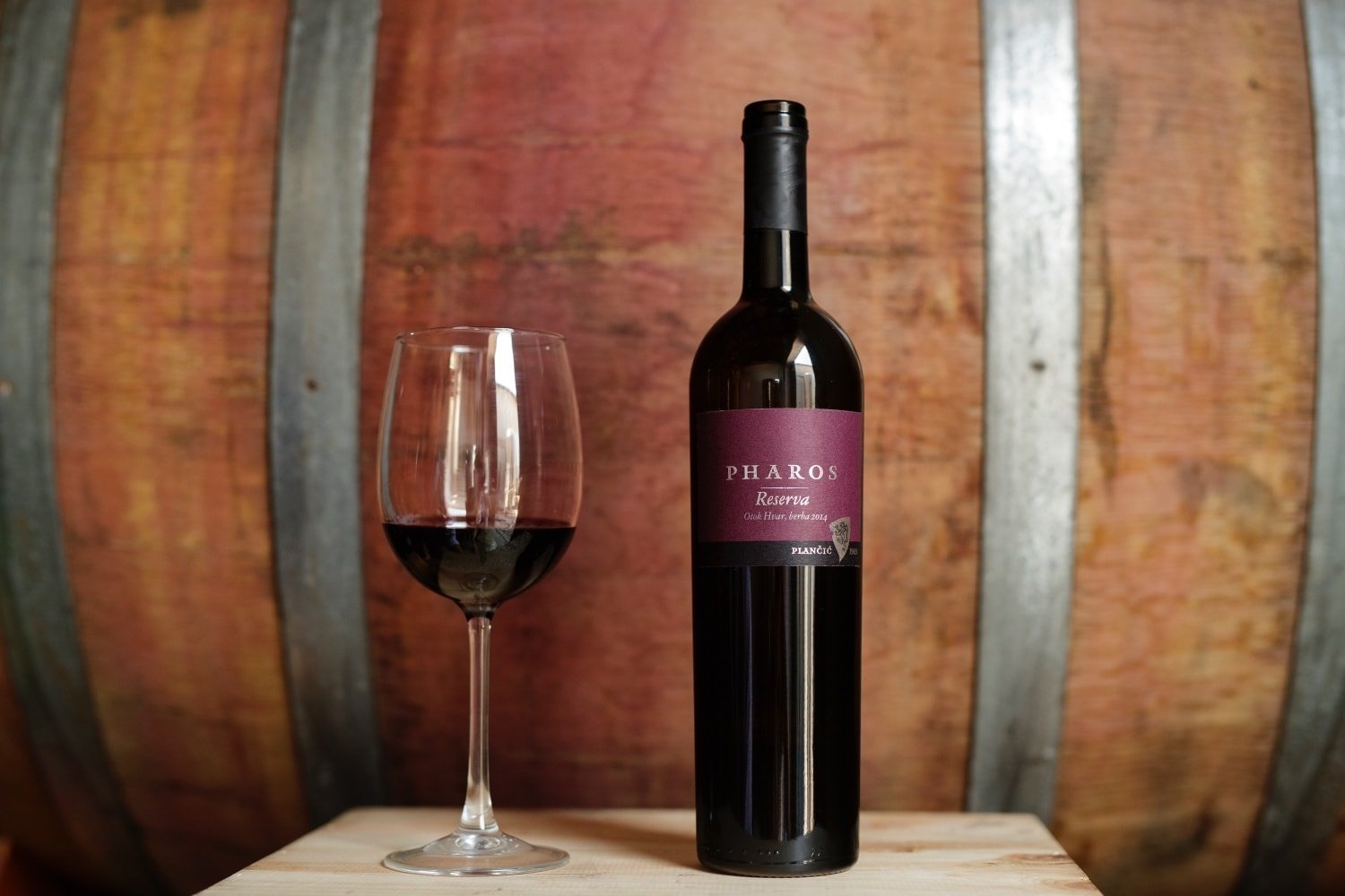 This image shows a bottle of Pharos Reserva wine and a glass filled with red wine. In the background, a wooden wine barrel. 