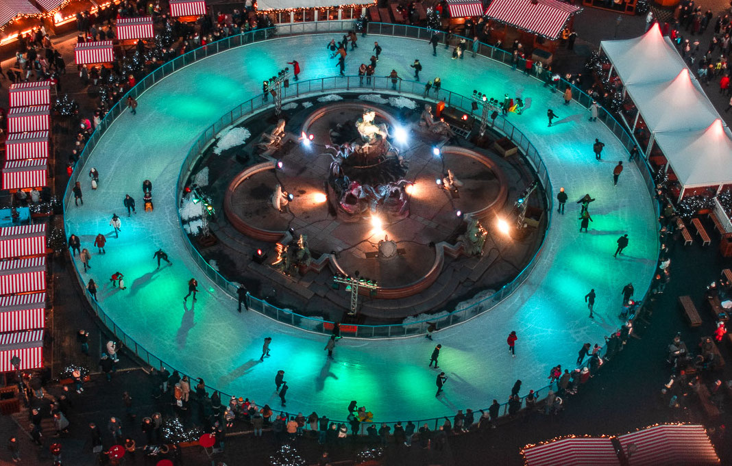 This is the image of an ice-skating rink from above. The skating rink is situated in the heart of a Christmas market. There are quite a few people skating and a lot more watching. 