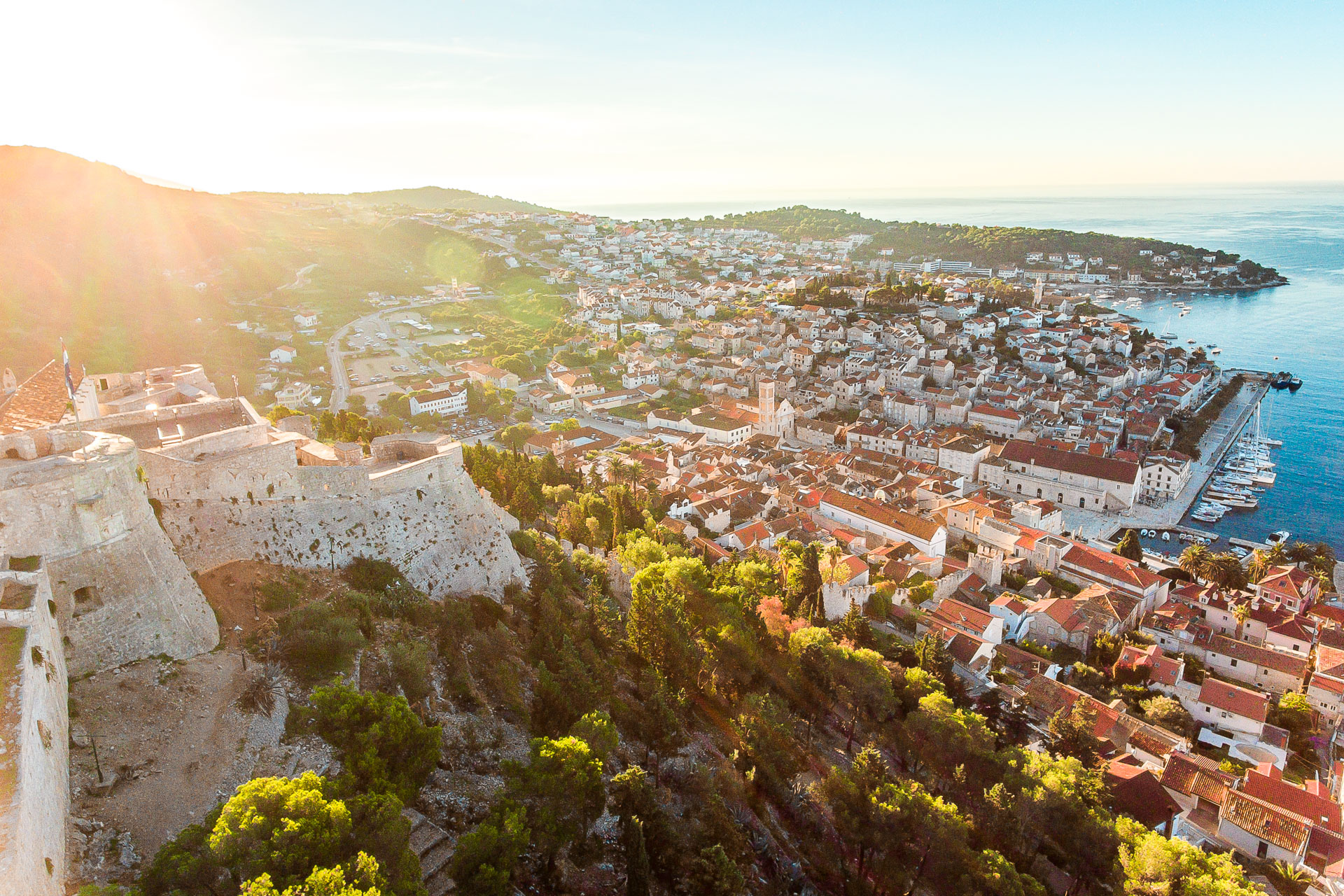 This is a panoramic shot of Hvar in Croatia. In the foreground, there's a castle that overlooks the red rooftops of the seaside town below. 